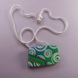 S342 This roughly rectangular pendant shades from blue to green with a white design. It has inset stripes of black and silver, green and gold. It is suspended on a 46cm (18 inches) sterling silver snake chain.