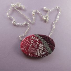 S343 An oval pendant in shades of red with a fine white design. It has a stripe of black and silver, and one end has a deep texture. It is suspended from a long diamond cut sterling silver chain, which measures 60cm (24 inches). S343 dark red black and silver silk screened pendant
